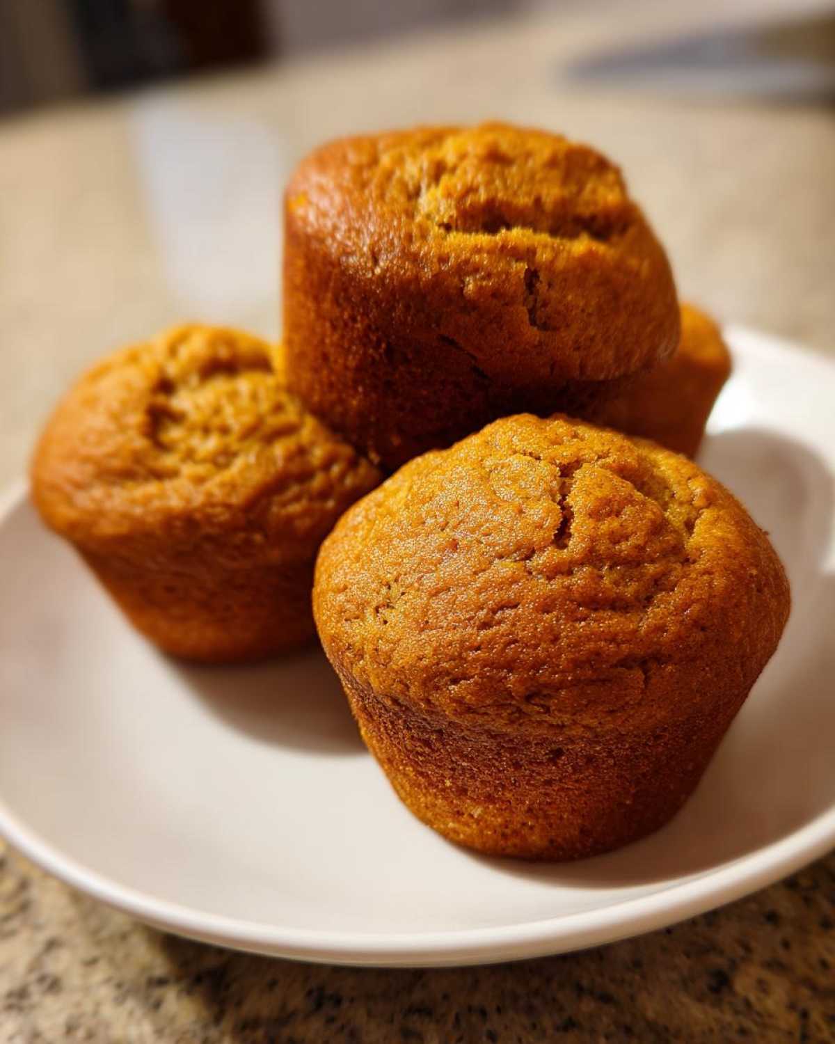 A close-up of four golden-brown banana protein muffins stacked slightly on a white plate.