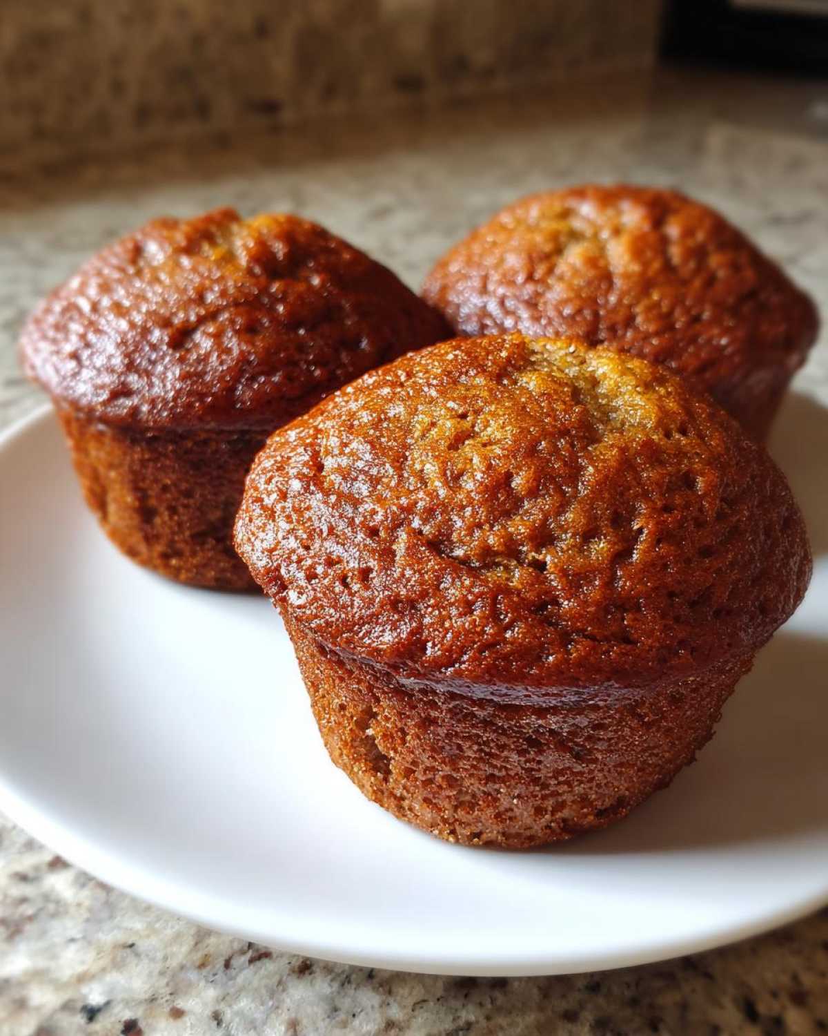 Three freshly baked, golden-brown banana protein muffins sitting closely together on a white plate.