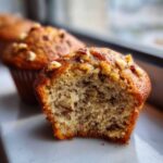 A close-up view of a banana nut muffin with a bite taken out, showing the moist interior and chopped nuts.