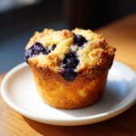 A single, golden-brown blueberry oatmeal muffin with a sugary crumb topping, sitting on a small white plate.