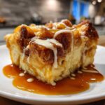 A close-up of a square serving of apple bread pudding drizzled heavily with caramel sauce and vanilla glaze.