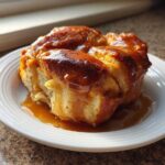 A single, golden brown apple dumpling covered in shiny caramel sauce, served on a white plate.