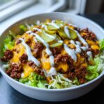 A close-up of a hearty cheeseburger bowl featuring seasoned ground beef, melted cheddar cheese, shredded lettuce, and pickle slices drizzled with white sauce.