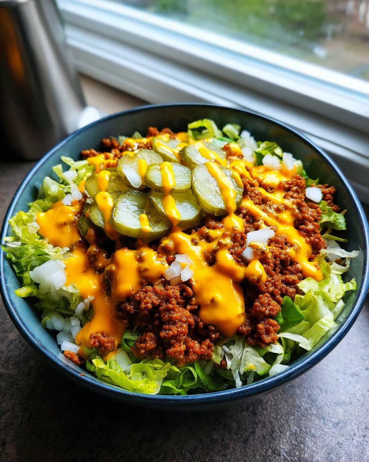 A close-up of a vibrant cheeseburger bowl featuring seasoned ground beef, shredded lettuce, cheese sauce, and pickle slices.