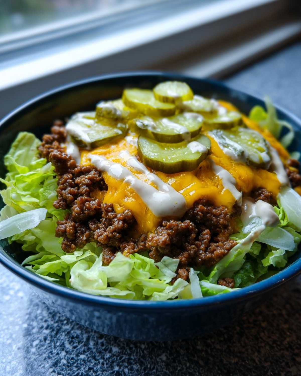 A close-up of a cheeseburger bowl featuring seasoned ground beef, melted cheddar cheese, shredded lettuce, pickles, and a drizzle of sauce.