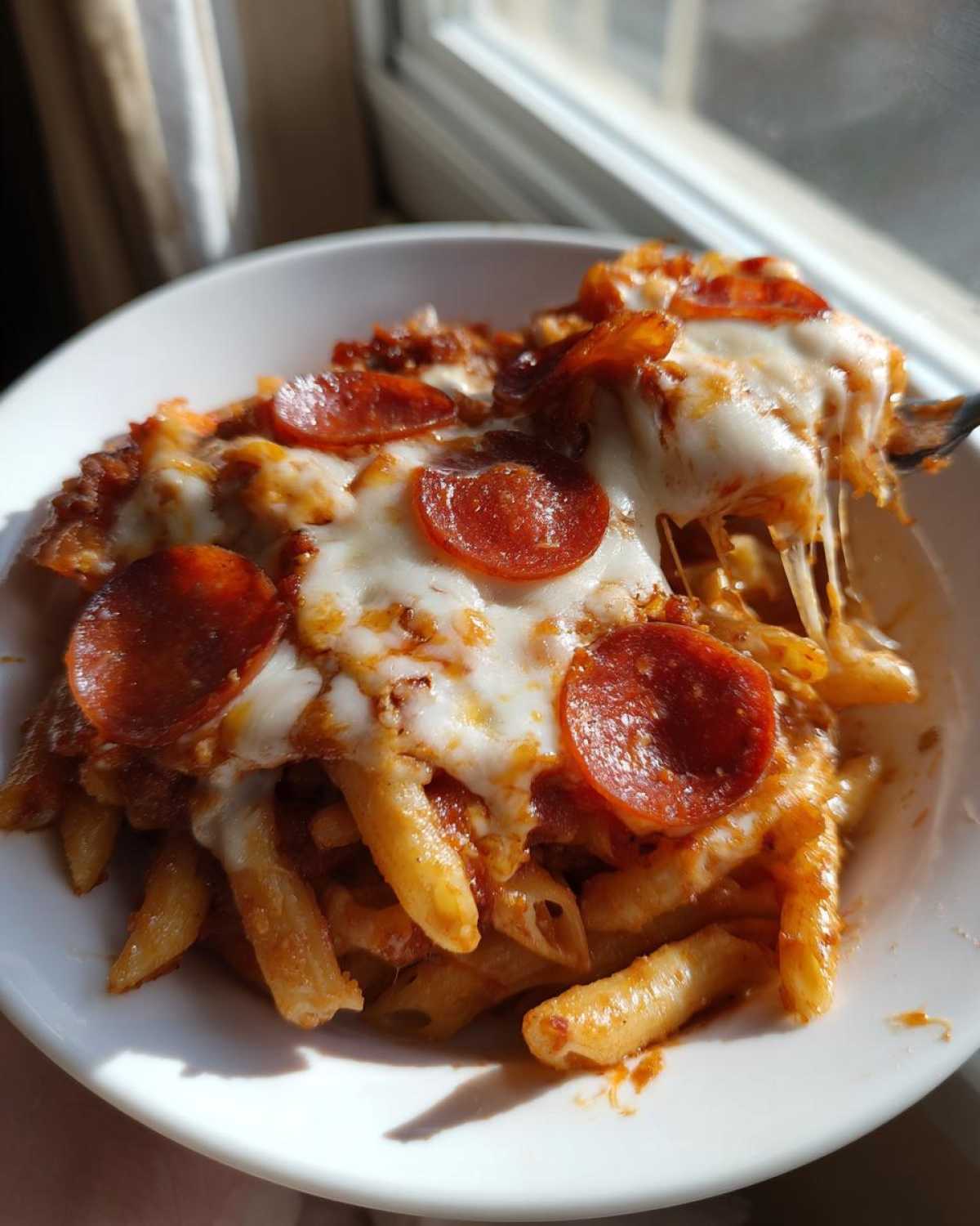 A serving of cheesy pizza pasta bake with melted mozzarella and pepperoni being lifted by a fork.