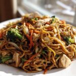 Close-up of a generous serving of chicken chow mein recipe with noodles, chicken pieces, broccoli, and carrots on a white plate.