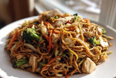 Close-up of a generous serving of chicken chow mein recipe with noodles, chicken pieces, broccoli, and carrots on a white plate.
