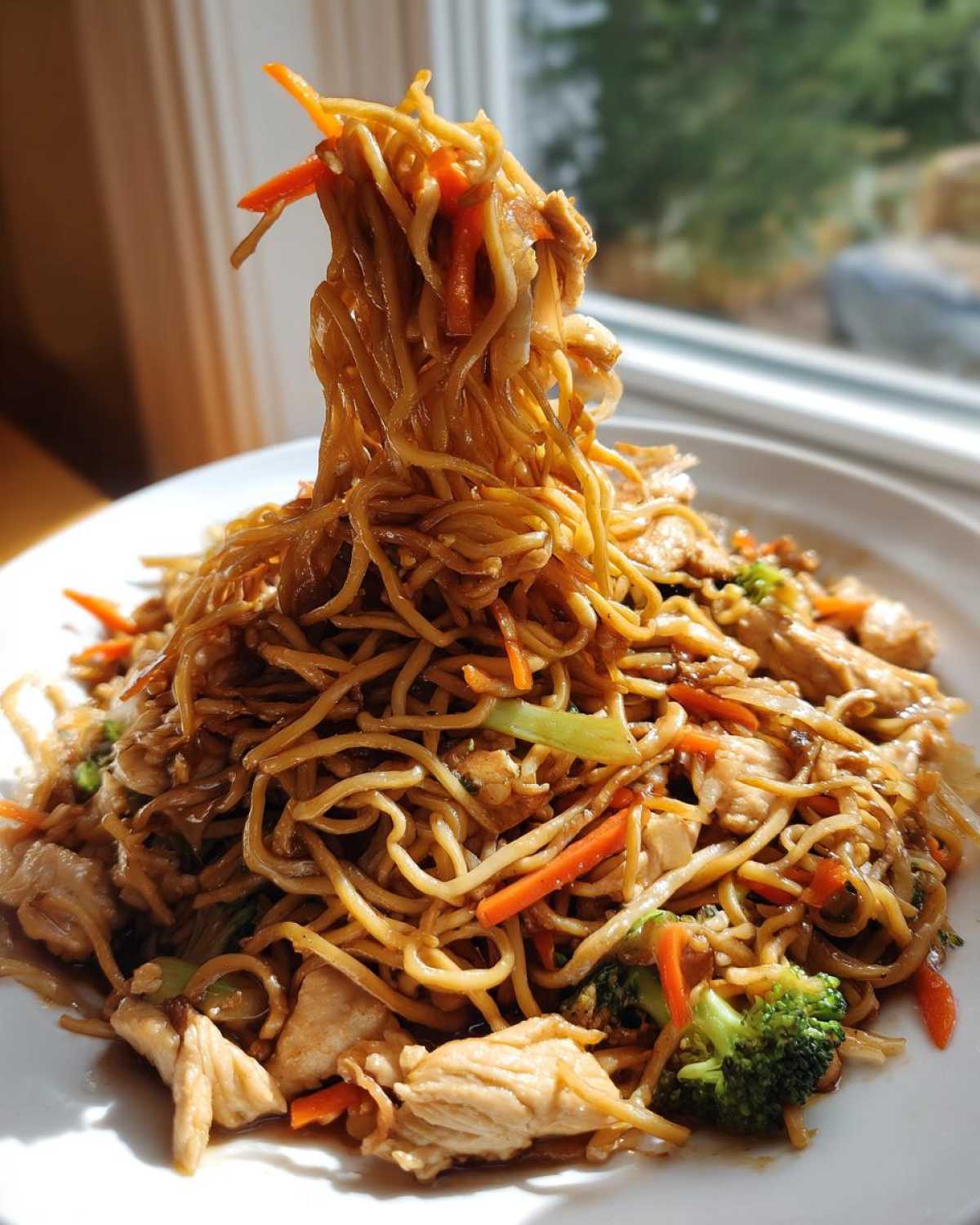 A fork lifts a large tangle of noodles from a plate of chicken chow mein recipe, showing chicken, carrots, and broccoli.