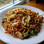 A close-up of a generous serving of chicken chow mein recipe featuring noodles, chicken pieces, broccoli, carrots, and cabbage.
