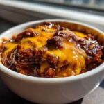Close-up of rich, dark chili topped with perfectly melted bright orange cheddar cheese in a white bowl, showcasing the chili cheese dip.