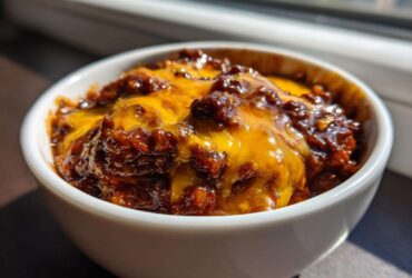 Close-up of rich, dark chili topped with perfectly melted bright orange cheddar cheese in a white bowl, showcasing the chili cheese dip.