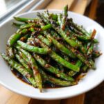 A white bowl filled with vibrant Chinese green beans with garlic sauce, showing blistered edges and glossy coating.