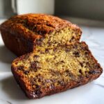 Close-up of a slice of chocolate chip vegan banana bread next to the loaf on a white marble surface.