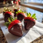 Close-up of freshly made chocolate covered strawberries with bright red tops and dark chocolate coating, resting on white parchment paper.