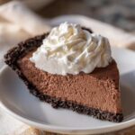 A close-up of a rich slice of chocolate mousse pie with a dark crust, topped with a swirl of fresh whipped cream.