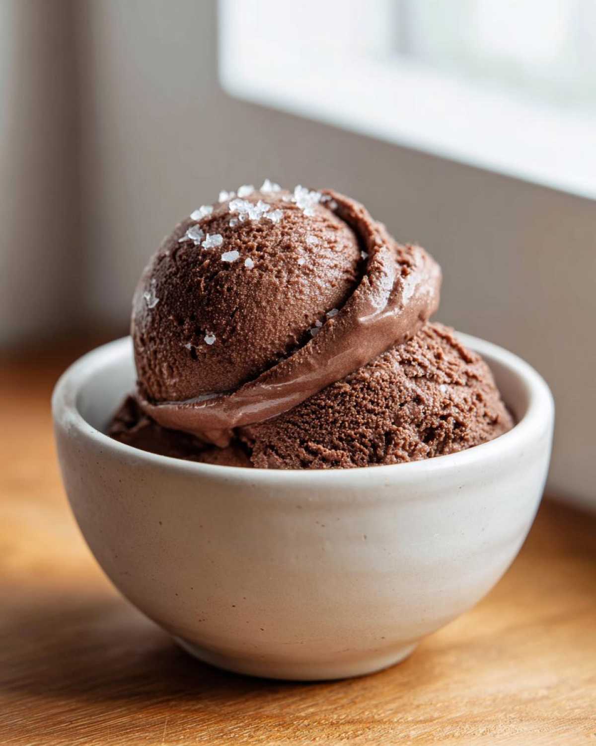 Two scoops of rich chocolate protein ice cream topped with flaky sea salt in a white bowl.