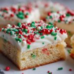 A close-up of a square slice of Christmas sugar cookie bars topped with thick white frosting and red and green holiday sprinkles.
