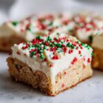 A single square of rich christmas sugar cookie bars topped with thick white frosting and festive red, green, and white sprinkles.