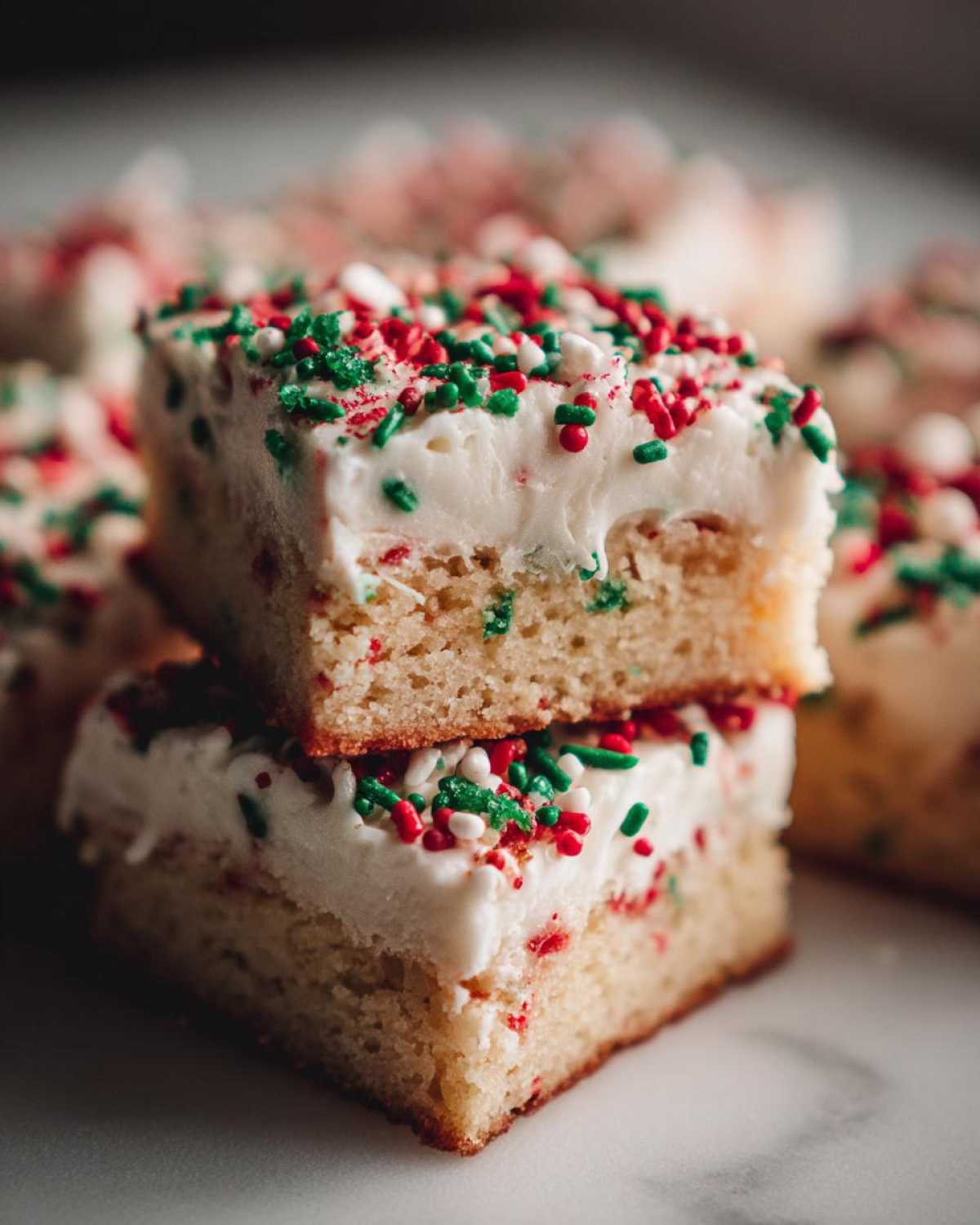 Two stacked squares of soft christmas sugar cookie bars topped with thick white frosting and red and green sprinkles.