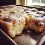 A close-up of a freshly baked cinnamon roll casserole in a pan, generously covered in dripping vanilla icing.