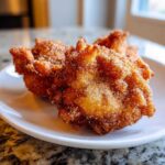 Three golden brown, irregularly shaped buñuelos heavily coated in cinnamon sugar, resting on a white plate.