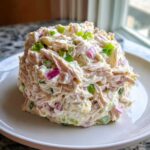 A mound of creamy cottage cheese chicken salad mixed with shredded chicken, red onion, and green onions on a white plate.