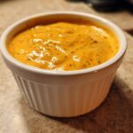 A close-up of smooth, orange beer cheese dip served in a small white ramekin on a speckled countertop.