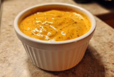 A close-up of smooth, orange beer cheese dip served in a small white ramekin on a speckled countertop.