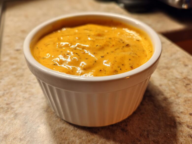 A close-up of smooth, orange beer cheese dip served in a small white ramekin on a speckled countertop.