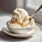 A close-up of a generous scoop of creamy, pale cottage cheese ice cream in a white bowl, with a silver spoon dipped in.
