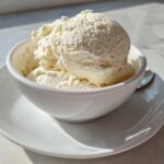 Two scoops of creamy, white cottage cheese ice cream served in a small white bowl on a matching saucer.