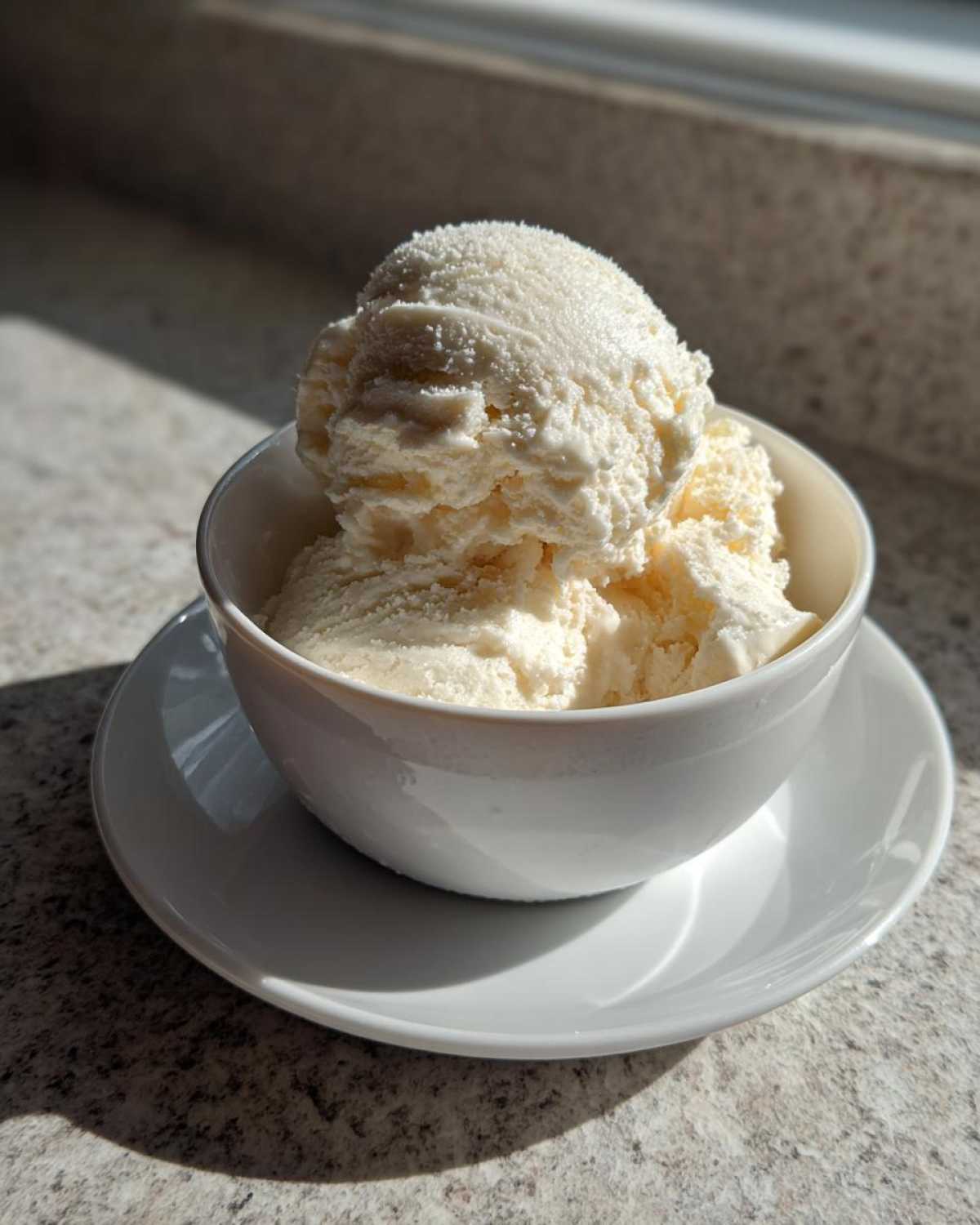 Two scoops of creamy, vanilla-colored cottage cheese ice cream served in a white bowl on a saucer.