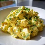 A close-up of creamy, yellow egg salad recipe mixed with green herbs, served on a white plate.