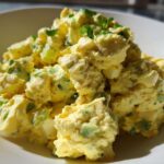 A close-up, brightly lit serving of creamy egg salad recipe mixed with green celery and herbs in a white bowl.