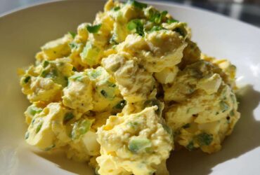 A close-up, brightly lit serving of creamy egg salad recipe mixed with green celery and herbs in a white bowl.