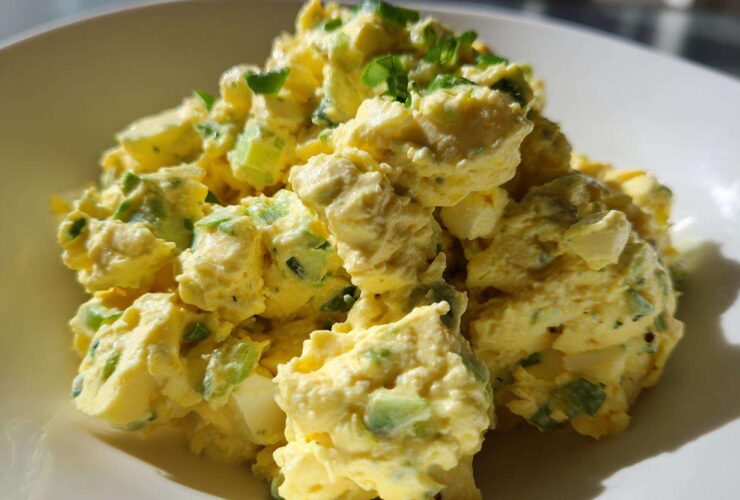 A close-up, brightly lit serving of creamy egg salad recipe mixed with green celery and herbs in a white bowl.