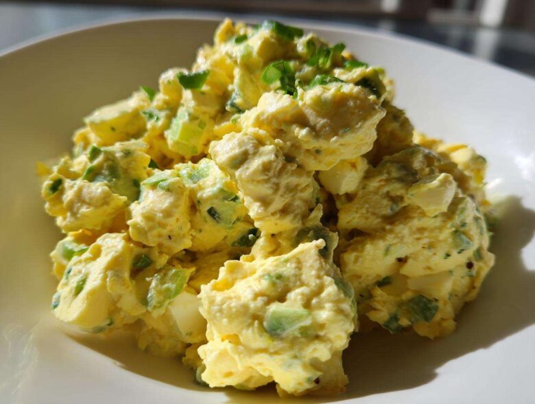 A close-up, brightly lit serving of creamy egg salad recipe mixed with green celery and herbs in a white bowl.