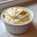 A small white ramekin filled with thick, creamy homemade garlic aioli sitting on a light countertop.