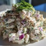 A close-up, bright photo of creamy greek yogurt chicken salad mixed with chunks of red onion and topped with fresh dill.
