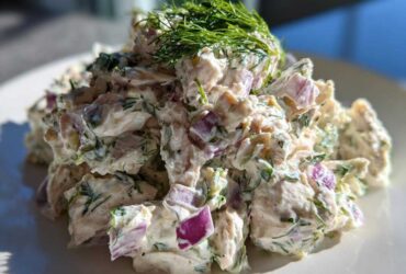 A close-up, bright photo of creamy greek yogurt chicken salad mixed with chunks of red onion and topped with fresh dill.