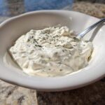 Close-up of a white bowl filled with thick, creamy horseradish sauce, garnished with dried herbs.