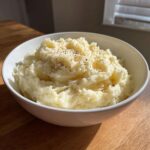 A close-up of a white bowl filled with creamy, fluffy mashed potatoes, topped generously with cracked black pepper.