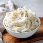 A close-up of a white bowl filled with fluffy, creamy mashed potatoes, topped generously with cracked black pepper.