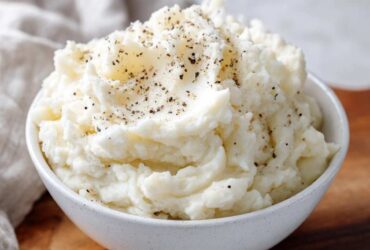 A close-up of a white bowl filled with fluffy, creamy mashed potatoes, topped generously with cracked black pepper.