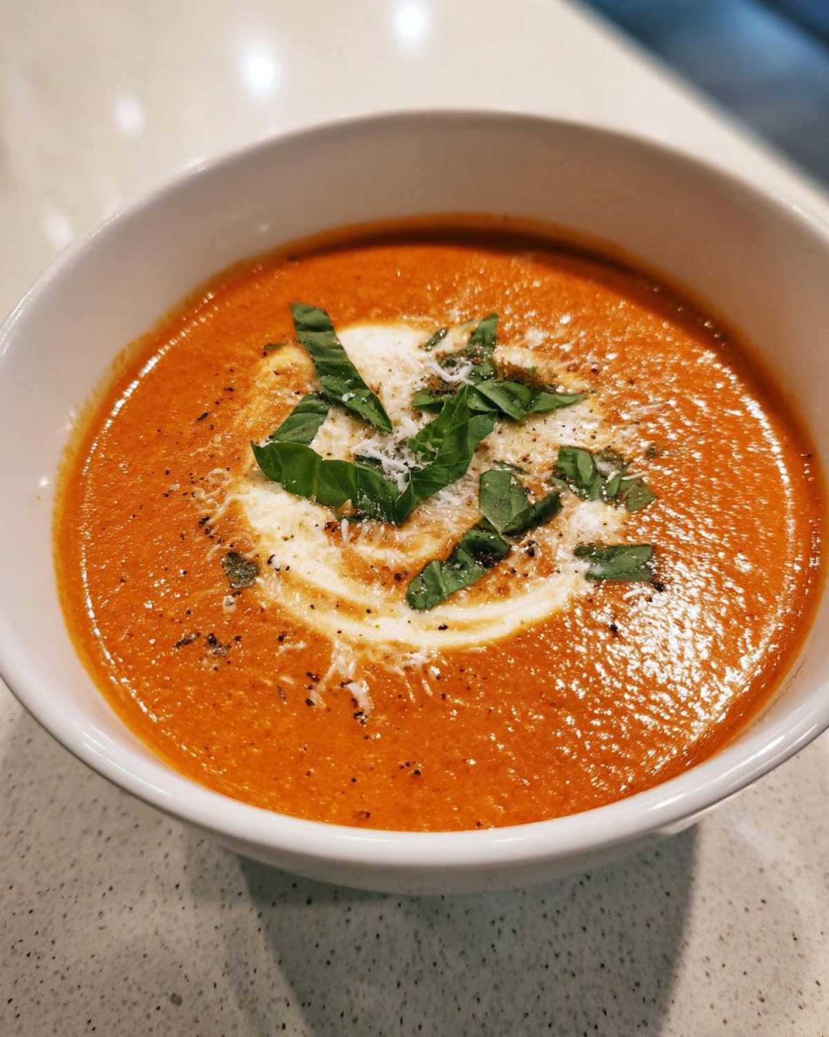 Close-up of a bowl of creamy tomato basil soup, garnished with a swirl of cream, fresh basil leaves, and grated cheese.