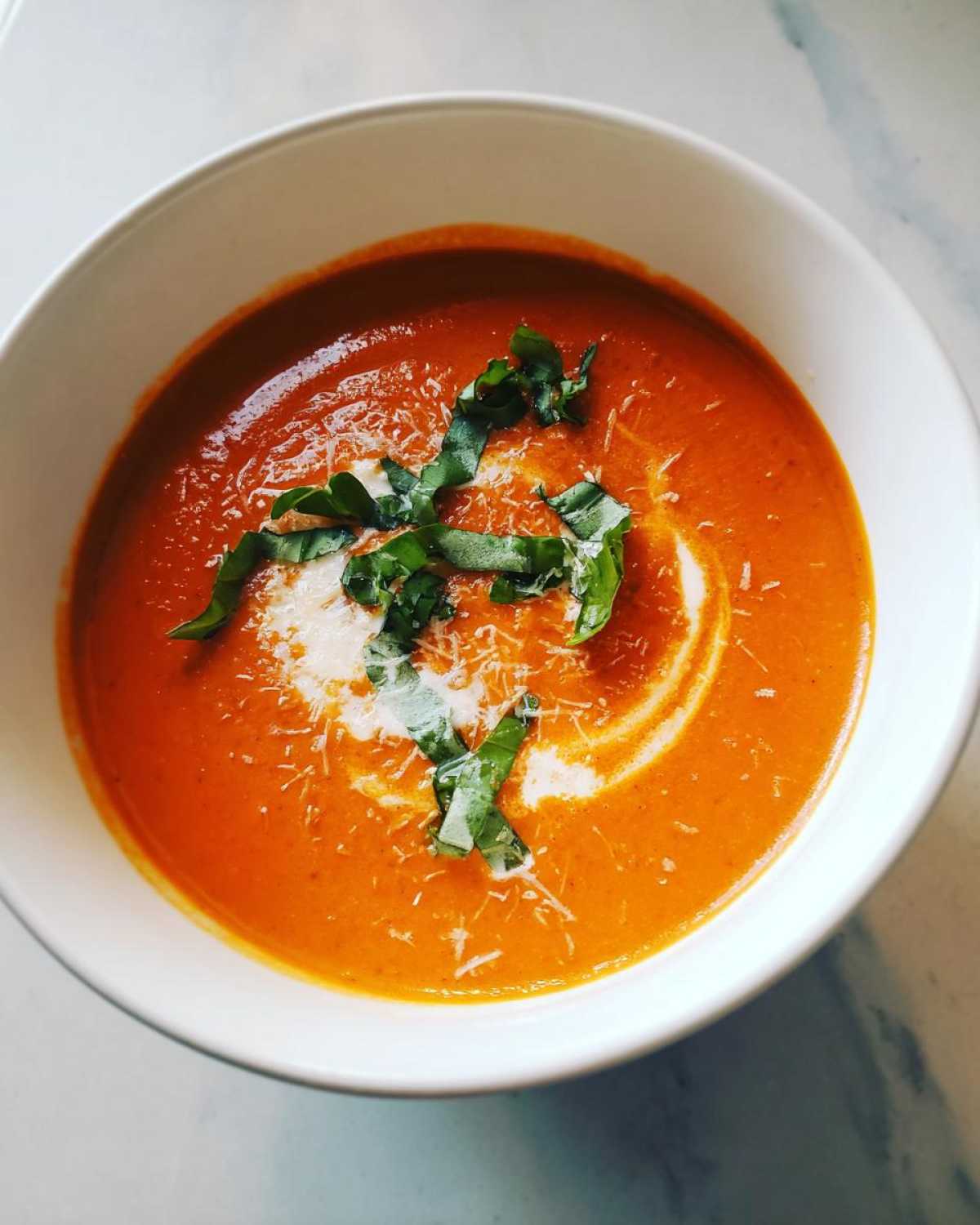 Overhead view of a bowl of creamy tomato basil soup, garnished with a swirl of cream, shredded cheese, and fresh basil leaves.