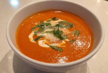 A close-up of a bowl of creamy tomato basil soup, garnished with basil and grated cheese.