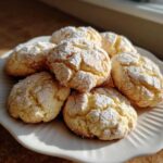 A pile of soft, crinkled, powdered sugar-dusted cool whip cookies resting on a white, scalloped plate.