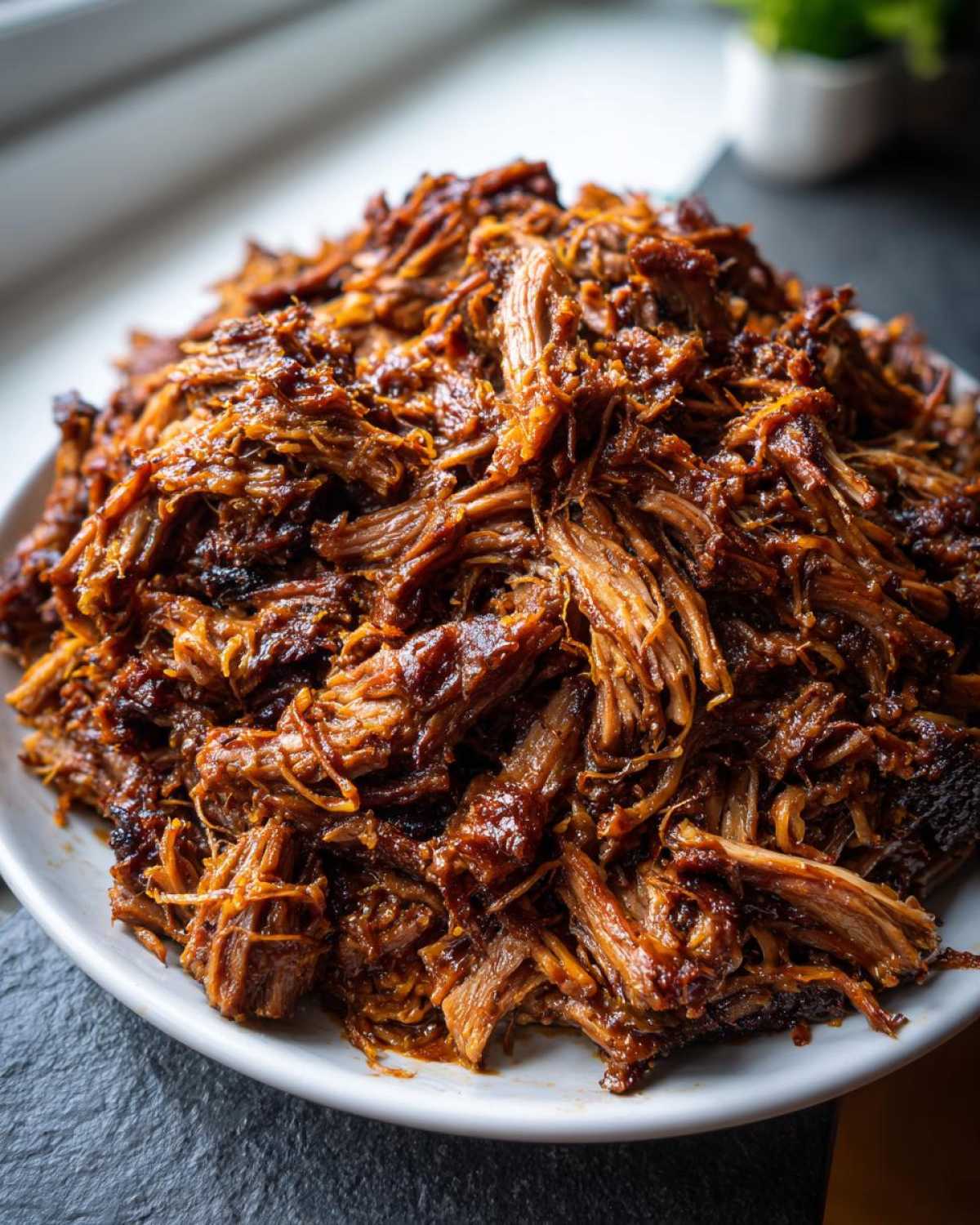 A close-up of a large mound of perfectly shredded, saucy crock pot pulled pork on a white plate.